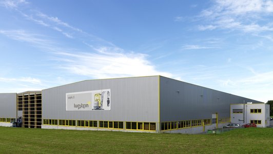 Außenansicht der Lugstein Fahrerkabinen Produktionshalle, mit grauer Fassade und gelben Akzenten, umgeben von einer Wiese