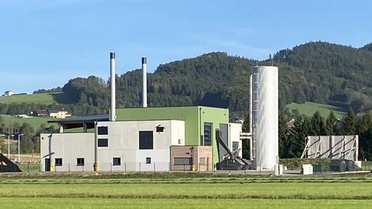 Nahwärmeanlage mit Holzlager und rauchendem Schornstein, klare Wetterbedingungen, umliegende Landschaft