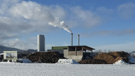Nahwärmeanlage mit Holzstapeln im Vordergrund und rauchenden Schornsteinen, schneebedeckter Boden