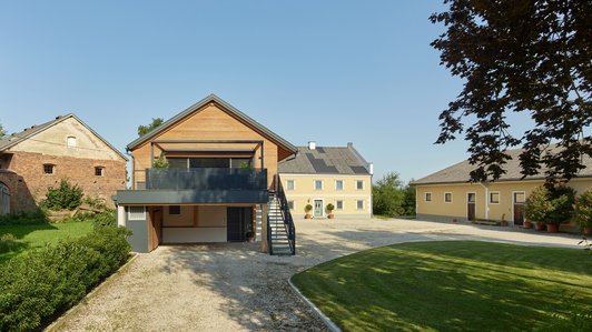 Ansicht eines weitläufigen Hofes mit einem modernen Holzhaus im Vordergrund und traditionellen Gebäuden im Hintergrund