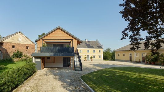 Ansicht eines weitläufigen Hofes mit einem modernen Holzhaus im Vordergrund und traditionellen Gebäuden im Hintergrund