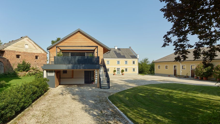 Ansicht eines weitläufigen Hofes mit einem modernen Holzhaus im Vordergrund und traditionellen Gebäuden im Hintergrund