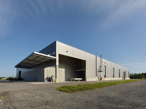 Weitwinkelansicht einer grauen Lagerhalle mit Nebengebäuden auf grünem Rasen, blauer Himmel im Hintergrund