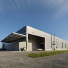 Weitwinkelansicht einer grauen Lagerhalle mit Nebengebäuden auf grünem Rasen, blauer Himmel im Hintergrund